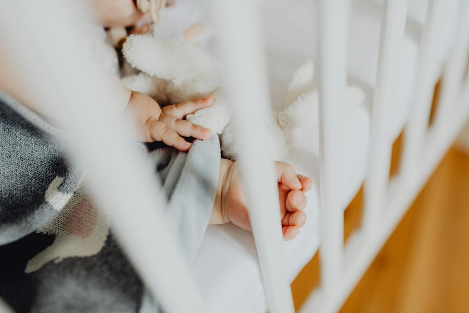 Tiny baby hands resting through the bars of a white crib beside a soft lamb toy